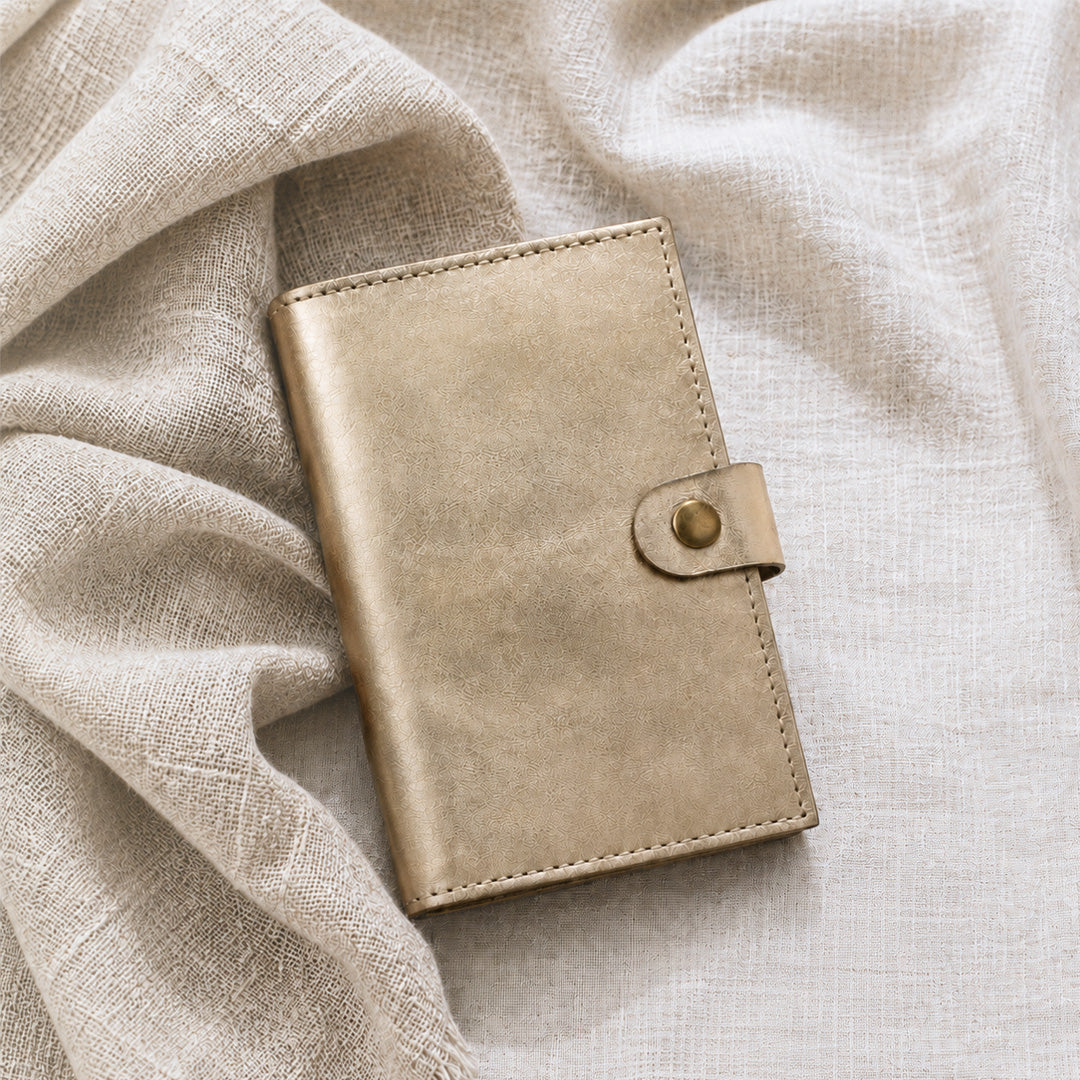 Cartera MANIKĀ de piel vacuna metalizada dorada cerrada, apoyada sobre tela de lino natural